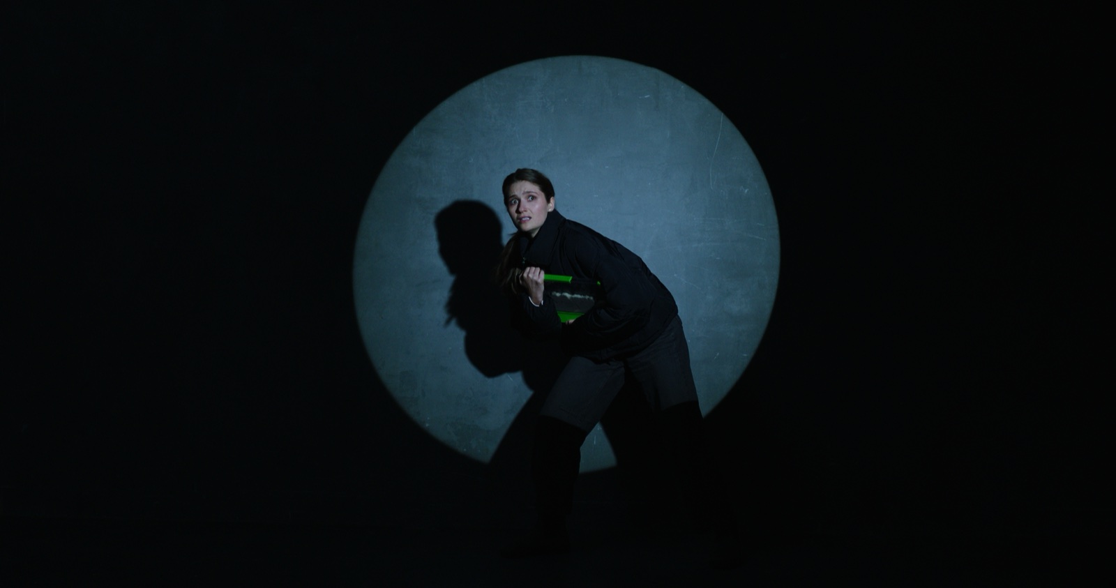 Still from I Can't Believe It's Not Better: a woman crouched in a spotlight against a dark stage wall.