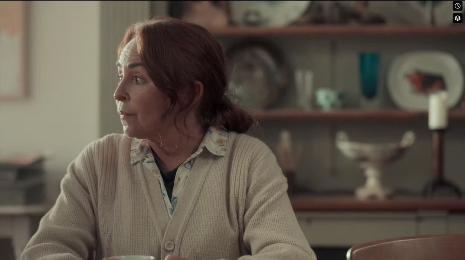 Still from Something Else: a woman in a cream cardigan at a table in a lived-in kitchen.